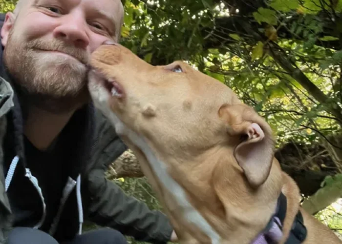 Man crouching outdoors while a tan dog wearing a harness licks his cheek, surrounded by green foliage and trees