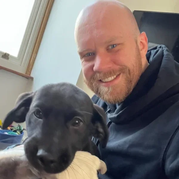 Man smiling indoors while holding a black puppy that is chewing a toy, with natural light from a nearby window