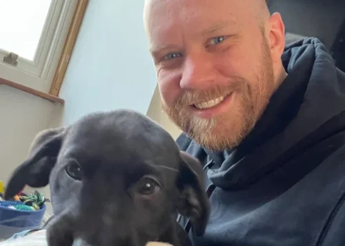Man smiling indoors while holding a black puppy that is chewing a toy, with natural light from a nearby window