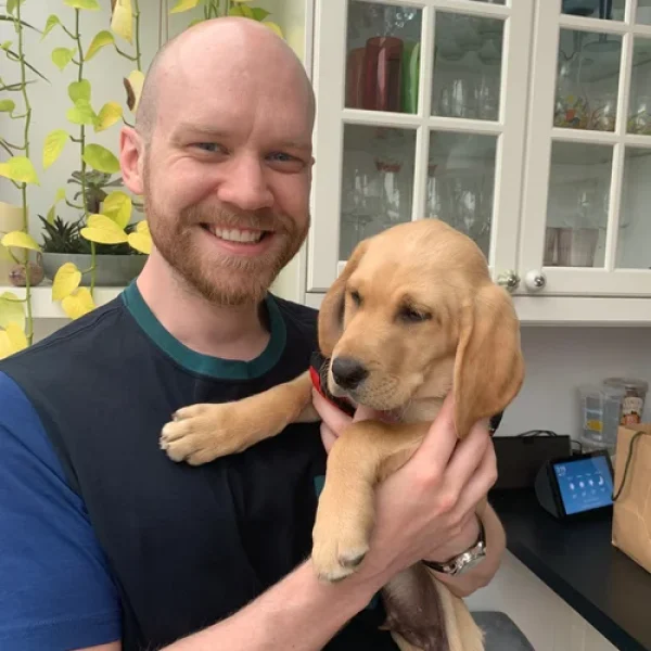 Man smiling in a kitchen while holding a Golden Retriever puppy, with houseplants and glass cabinet in the background